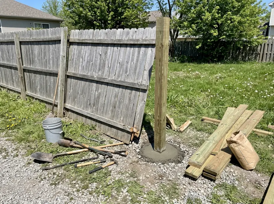 A weathered wood fence section leaning after wind, with a new post set in concrete beside the broken one
