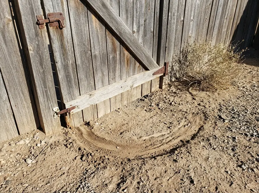A wood yard gate sagging on its hinges, its latch corner scraping a groove in the dirt