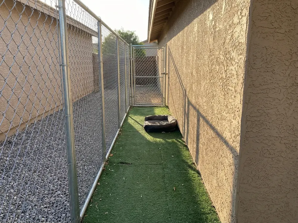 A long narrow chain link dog run along the side of a stucco house with artificial turf